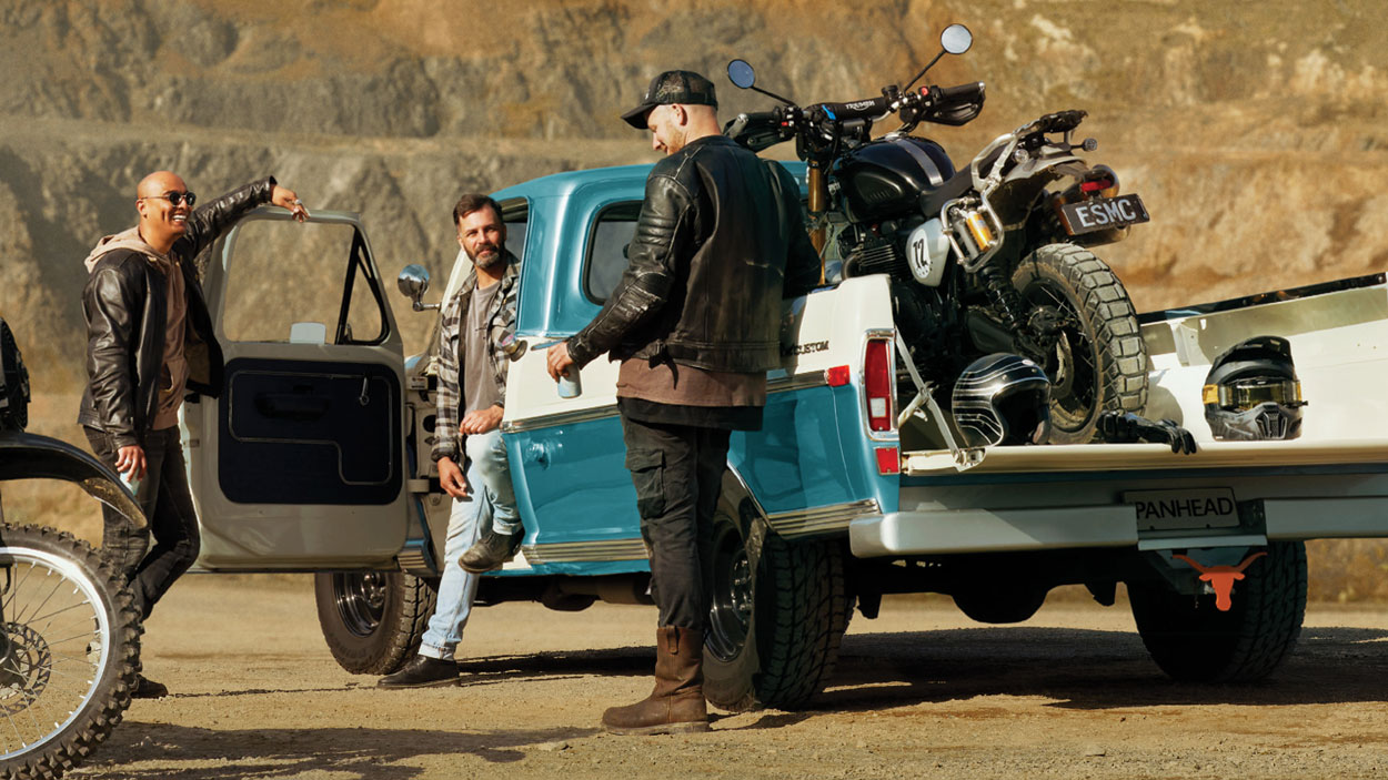 Photo of three people standing around a vintage pick-up truck in the desert, with dirt bikes